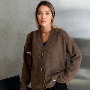 May include: A brown, button-down cardigan sweater with a V-neck and two front pockets. The sweater has three visible buttons and is made of a soft, knitted material. A brown shoulder bag is visible on the left side.