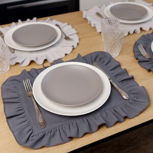 May include: A dining table setting with gray plates, white plates, and ruffled placemats. Silverware, including forks and knives, is arranged around the plates. The placemats and plates are in shades of gray and white.