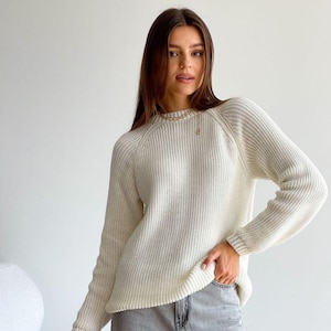 May include: Cream-colored knit sweater with long sleeves and a crew neck. The sweater has a ribbed texture and a small emblem near the neckline. The model is wearing gray jeans. The sweater is a casual, everyday garment.