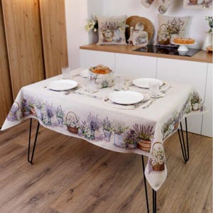 May include: A rectangular tablecloth with a lavender floral design, set on a table with black hairpin legs. The table is set with white plates, glasses, and placemats. Matching pillows and kitchen accessories are visible in the background.