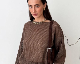 May include: A woman wearing a brown, open-weave sweater with long sleeves. She is also wearing a gold chain necklace and earrings. A brown suede shoulder bag with a silver buckle is also visible.