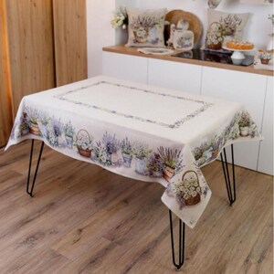 May include: A rectangular tablecloth with a floral lavender design. The off-white cloth features a border of lavender plants in pots and baskets. The table has black hairpin legs and is set on a wooden floor.