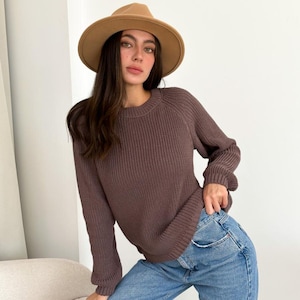 May include: A brown sweater with a crew neck and long sleeves. The sweater is paired with blue jeans and a tan fedora hat. The outfit is casual and suitable for fall or winter.