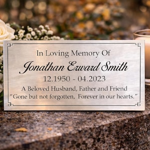 Könnte beinhalten: Rechteckige Gedenktafel mit der Inschrift "In Loving Memory Of Jonathan Erward Smith 12.1950 - 04.2023". Die Tafel befindet sich in der Nähe von Blumen und einer brennenden Kerze, was eine ergreifende Szene der Erinnerung schafft.
