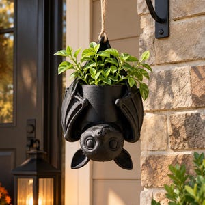 May include: A black hanging planter shaped like a bat suspended by a jute rope from a metal wall bracket. The bat is depicted in an upside-down position with its wings wrapped around a central cylindrical pot containing a small green plant.