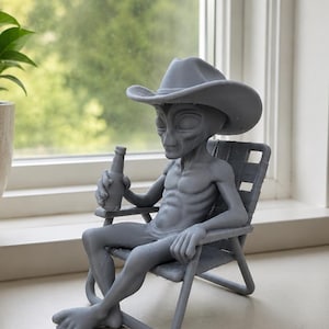 May include: A gray figurine of an extraterrestrial being wearing a cowboy hat, sitting in a folding lawn chair while holding a small bottle. The figure has a slender build, large oval eyes, and is depicted in a relaxed, seated posture.