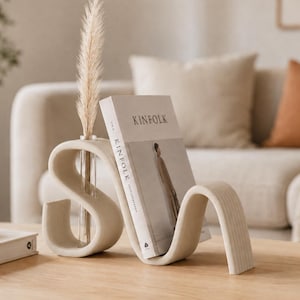 May include: A beige, S-shaped ceramic bookend and vase combination with a smooth, curved design. A clear glass tube is inserted into the top of the structure, holding a single dried pampas grass stem. The piece supports a copy of Kinfolk magazine.