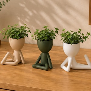 May include: Three ceramic planters shaped like human figures in beige, dark green, and white. Each planter features a rounded head that holds a small green ivy plant, with the figure's body forming the base and legs in various seated poses.