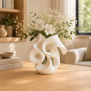 May include: A white ceramic vase with a sculptural, swirling, organic form featuring multiple open loops and curves. The vase holds a large, airy arrangement of white baby's breath flowers and green foliage, creating a textured, layered appearance.
