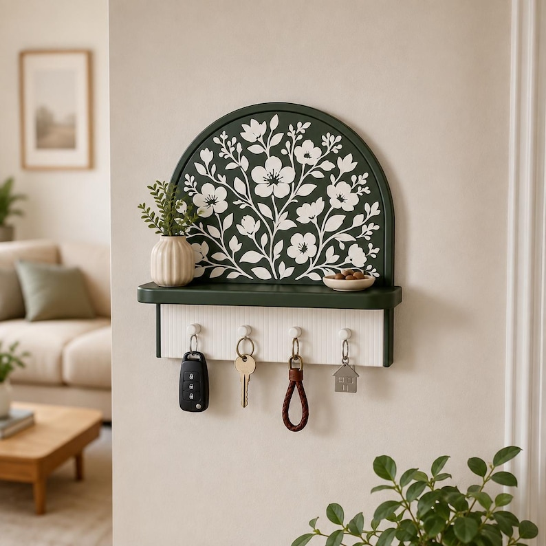 Puede incluir: Un soporte para llaves de pared en color verde oscuro con un respaldo arqueado que presenta un patr&oacute;n floral blanco. La unidad incluye un peque&ntilde;o estante con un jarr&oacute;n acanalado y un cuenco, adem&aacute;s de cuatro ganchos blancos con llaves colgadas.