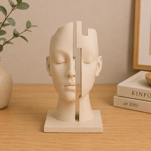 May include: A beige sculpture of a human head split vertically into two separate pieces. The sculpture features closed eyes, a smooth matte finish, and a square base. The two halves are positioned slightly apart, revealing a gap between them.