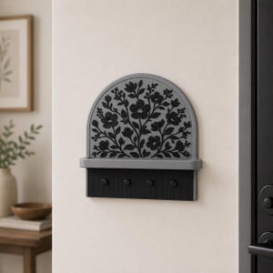 Puede incluir: Un soporte para llaves de pared con una parte superior arqueada gris y un patr&oacute;n floral negro en relieve. Debajo del arco hay un peque&ntilde;o estante horizontal y una base rectangular negra con cuatro ganchos circulares para colgar objetos.