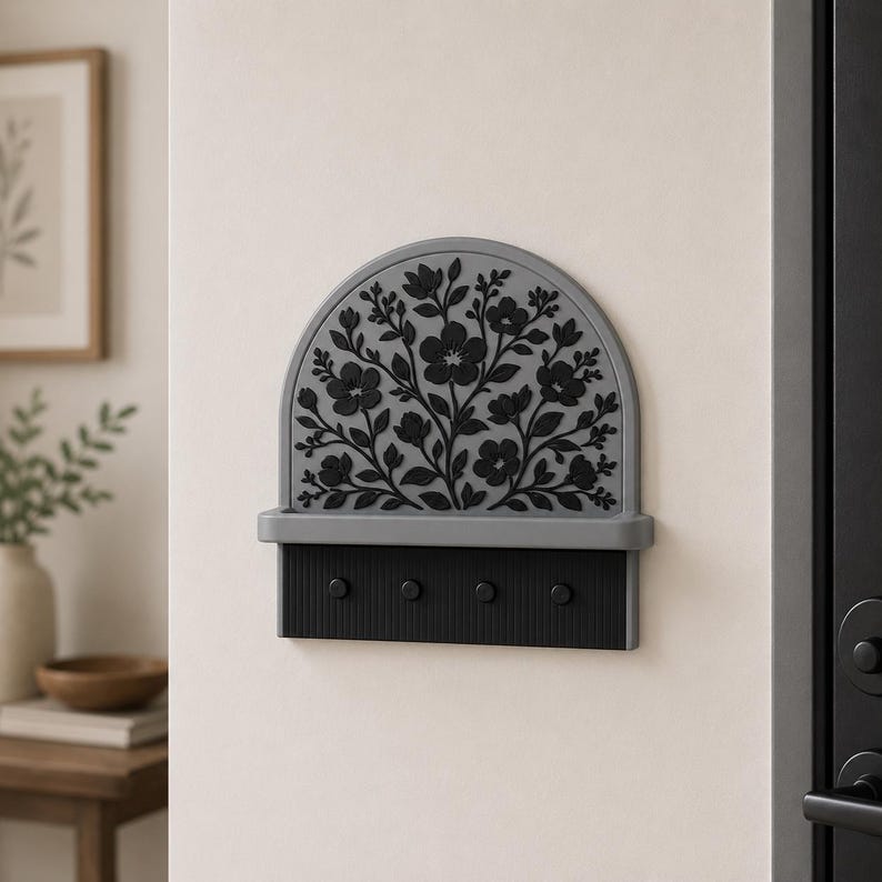 Puede incluir: Un soporte para llaves de pared con una parte superior arqueada gris y un patr&oacute;n floral negro en relieve. Debajo del arco hay un peque&ntilde;o estante horizontal y una base rectangular negra con cuatro ganchos circulares para colgar objetos.