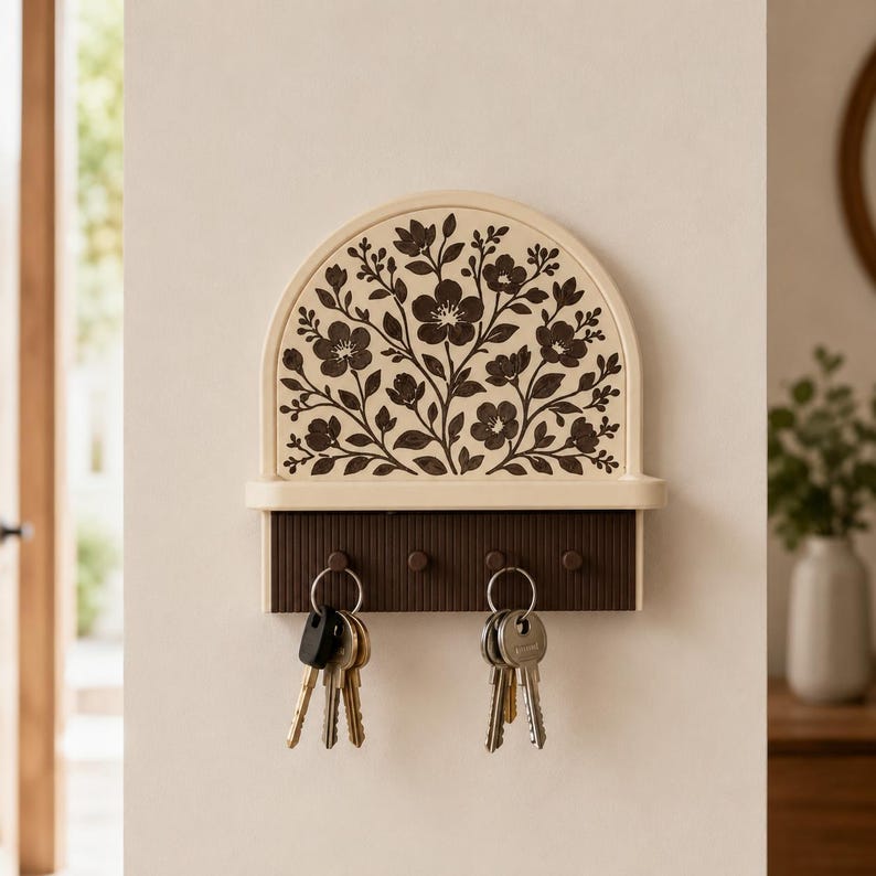 Puede incluir: Un soporte para llaves de pared con una parte superior arqueada que presenta un patr&oacute;n floral marr&oacute;n oscuro sobre un fondo crema. La unidad incluye un peque&ntilde;o estante y una base acanalada marr&oacute;n oscuro con cuatro ganchos, dos con llaves.