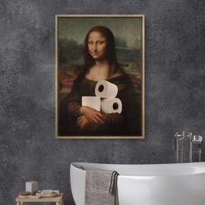 May include: Framed print of the Mona Lisa holding rolls of toilet paper. The artwork is in a wooden frame and hangs on a dark gray wall. The bathroom setting includes a white bathtub and a small wooden stool.