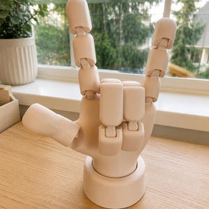 May include: A light beige articulated hand model made of smooth material, featuring a round base and movable joints. The fingers are positioned in a rock-and-roll gesture with the index and pinky fingers extended upward while the middle fingers are folded.