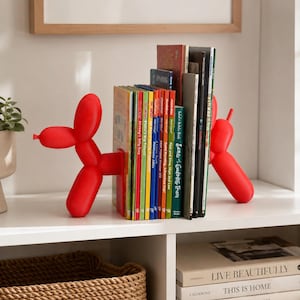 May include: A pair of bright red balloon dog bookends made of a matte material, positioned on opposite sides of a row of colorful children's books. The bookends feature a stylized, segmented design resembling a balloon animal with a snout and tail.