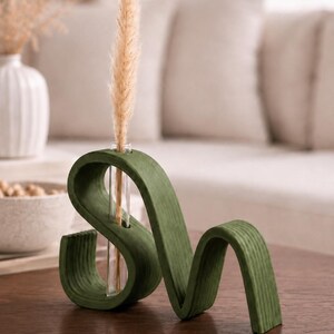 May include: A dark green, wavy, S-shaped wooden stand with a clear glass test tube inserted into the center. A single, tall, beige dried grass stem is placed inside the glass tube, extending upwards from the center of the curved wooden structure.