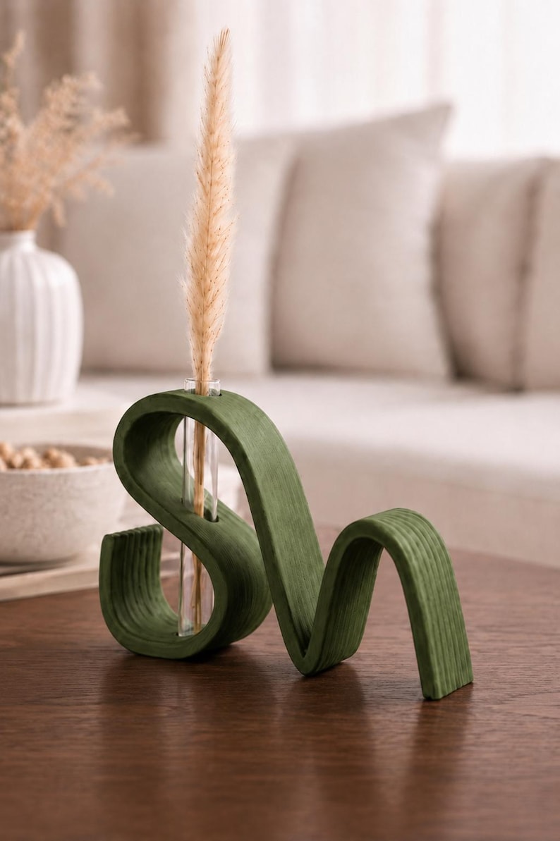 May include: A dark green, wavy, S-shaped wooden stand with a clear glass test tube inserted into the center. A single, tall, beige dried grass stem is placed inside the glass tube, extending upwards from the center of the curved wooden structure.