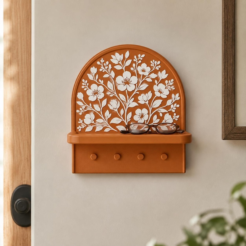 Puede incluir: Un estante de pared arqueado en acabado naranja quemado con un patr&oacute;n floral blanco en relieve. El estante incluye una repisa plana que sostiene unas gafas de montura oscura y una secci&oacute;n inferior con cuatro peque&ntilde;os ganchos circulares.