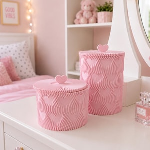 May include: Two cylindrical pink storage containers with lids, each topped with a small heart-shaped handle. The exterior of both containers features a textured, wavy pattern interspersed with heart shapes, creating a uniform, tactile surface.