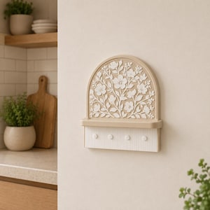 Puede incluir: Un soporte para llaves de pared con una parte superior arqueada que presenta un relieve floral blanco sobre un fondo color canela. Debajo del arco hay un peque&ntilde;o estante horizontal y una base rectangular blanca con cuatro ganchos circulares.