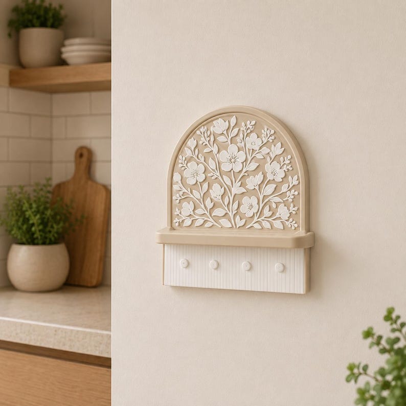 Puede incluir: Un soporte para llaves de pared con una parte superior arqueada que presenta un relieve floral blanco sobre un fondo color canela. Debajo del arco hay un peque&ntilde;o estante horizontal y una base rectangular blanca con cuatro ganchos circulares.