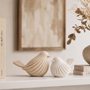 May include: Two bird figurines with a smooth, rounded shape and vertical grooved detailing. One bird is a light beige color, and the smaller bird is a crisp white. Both feature a pointed beak and a fanned tail, positioned side by side on a flat surface.