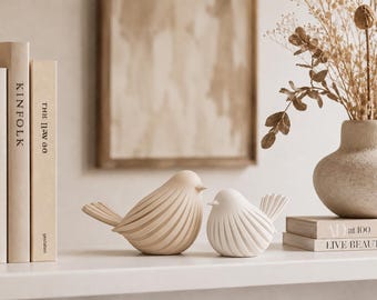May include: Two bird figurines, one in a light beige and one in a crisp white, both featuring a smooth, rounded body with deep, vertical carved grooves along the wings and chest, positioned side by side on a flat, level surface.
