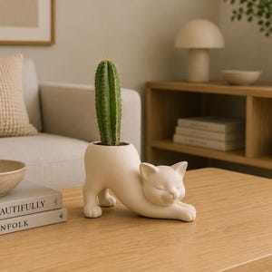May include: A matte off-white ceramic planter shaped like a cat in a stretching pose. The cat has its front paws extended forward and its back arched, with a green columnar cactus planted in the opening on its back. The surface is smooth.
