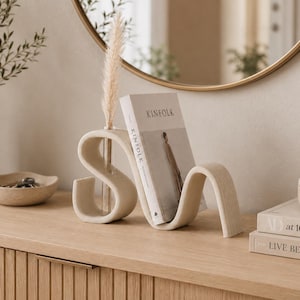 May include: A beige, S-shaped ceramic bookend with a built-in glass bud vase holding a dried pampas grass stem. The bookend supports a copy of Kinfolk magazine, featuring a neutral color palette and a smooth, matte finish on its curved structure.