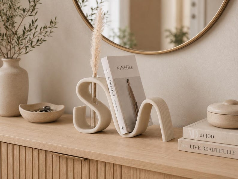 May include: A beige, S-shaped ceramic bookend with a built-in glass bud vase holding a dried pampas grass stem. The bookend supports a copy of Kinfolk magazine, featuring a neutral color palette and a smooth, matte finish on its curved structure.