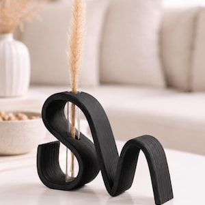 May include: A black wooden vase with a wavy, serpentine shape featuring a single clear glass test tube insert. A tall, slender, beige dried grass stem is placed inside the glass tube, extending upward from the center of the dark wood structure.