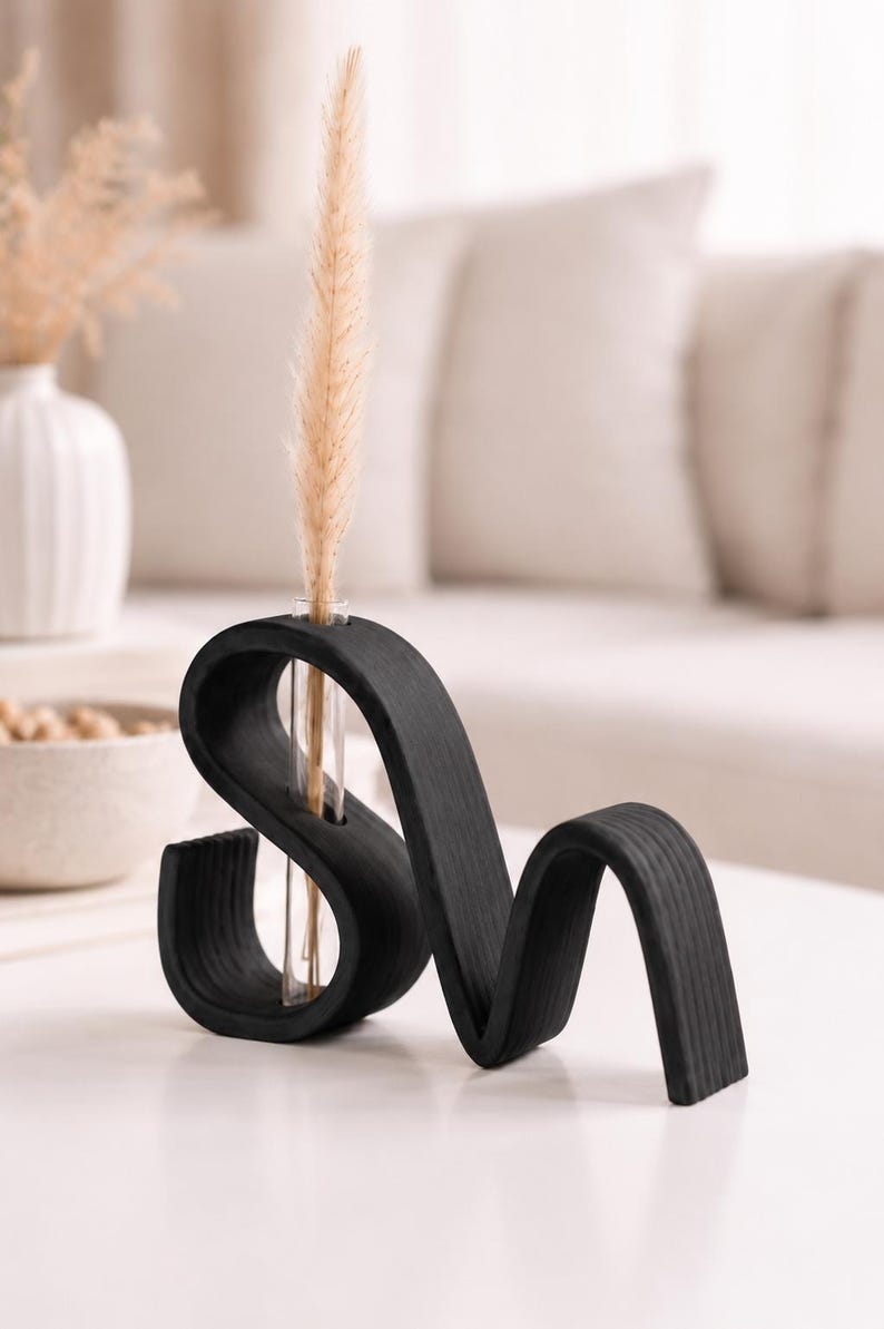 May include: A black wooden vase with a wavy, serpentine shape featuring a single clear glass test tube insert. A tall, slender, beige dried grass stem is placed inside the glass tube, extending upward from the center of the dark wood structure.