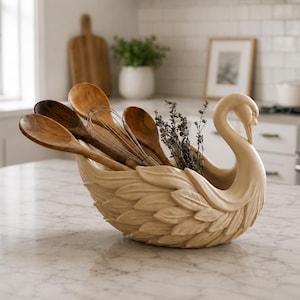 May include: A cream-colored ceramic swan-shaped bowl featuring detailed molded feather textures on its exterior. The bowl contains several wooden kitchen spoons, a metal wire whisk, and a small bundle of dried herbs tucked inside the basin.