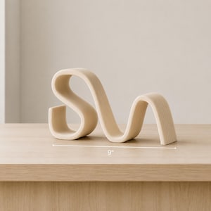 May include: A beige, wavy, sculptural object with a smooth, ribbed texture. The piece features a continuous, flowing form that curves into multiple loops. A measurement line indicates the object has a total width of 9 inches.
