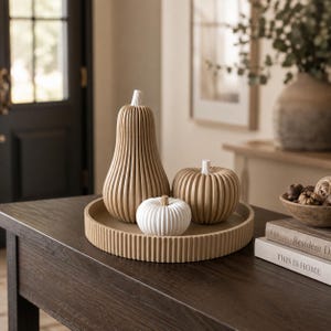 May include: A set of three ribbed decorative gourds and pumpkins in tan and white, arranged on a matching tan circular ribbed tray. The tallest gourd is tan, accompanied by a medium tan pumpkin and a smaller white pumpkin, all featuring vertical ridges.