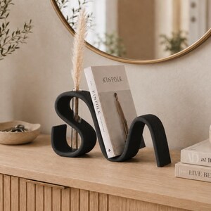 May include: A matte black, S-shaped sculptural bookend featuring a built-in clear glass test tube vase. The bookend holds a Kinfolk magazine upright, and a single dried pampas grass stem is placed inside the glass tube insert.