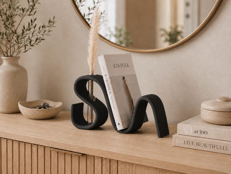 May include: A matte black, S-shaped sculptural bookend featuring a built-in clear glass test tube vase. The bookend holds a Kinfolk magazine upright, and a single dried pampas grass stem is placed inside the glass tube insert.