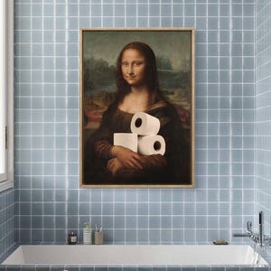 May include: Framed artwork of the Mona Lisa holding rolls of toilet paper, a humorous take on the classic painting. The artwork is displayed in a bathroom with blue tiled walls and a white bathtub.