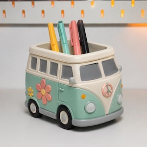 May include: A desk organizer shaped like a vintage van in pastel green and cream. The van features a pink flower decal on the side, a peace sign on the front, and gray bumpers. Four pens in yellow, teal, pink, and black are stored inside the top opening.