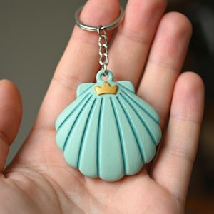 Könnte beinhalten: Ein hellblaues Muschel-Schlüsselanhänger mit goldenem Kronen-Akzent. Der Schlüsselanhänger besteht aus einem festen Material und hat eine silberne Kette und einen Schlüsselring. Das Muscheldesign weist detaillierte Linien und eine glatte Oberfläche auf.