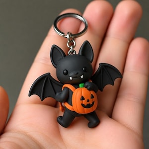 Könnte beinhalten: Ein schwarzer Fledermaus-Schlüsselanhänger mit matter Oberfläche, spitzen Ohren und zwei kleinen weißen Fangzähnen. Die Fledermaus trägt einen orangefarbenen, kürbisförmigen Rucksack mit einem geschnitzten Kürbisgesicht an einem silberfarbenen Ring.