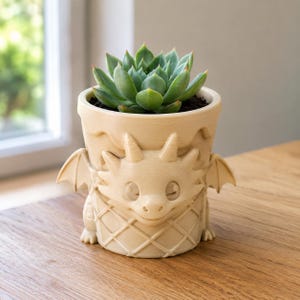 Op de afbeelding: Een lichtbeige, 3D-geprinte plantenpot in de vorm van een draak met kleine vleugels en een gestructureerd ruitpatroon aan de buitenkant. De pot bevat een groene vetplant met dikke, puntige bladeren in een strakke, ronde rozetvorm.