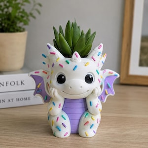 May include: A white dragon-shaped planter with a purple belly and wings, decorated with colorful sprinkles. The dragon has large black eyes, small horns, and its hands are resting under its chin. A green succulent plant is potted inside the top.