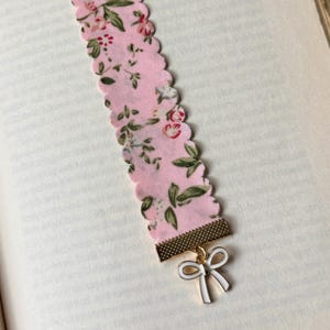 May include: A pink floral fabric bookmark with a scalloped edge. The bookmark has a gold-coloured metal piece at the bottom with a white bow charm. The fabric features a pattern of small flowers and green leaves.