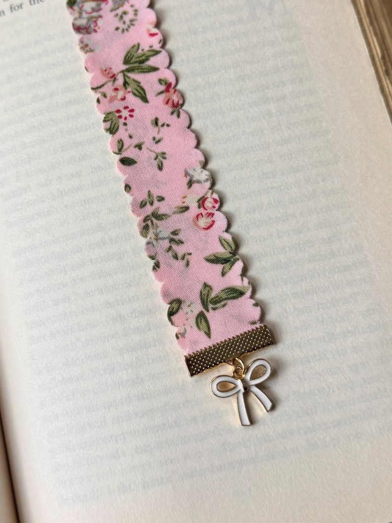 May include: A pink floral fabric bookmark with a scalloped edge. The bookmark has a gold-coloured metal piece at the bottom with a white bow charm. The fabric features a pattern of small flowers and green leaves.