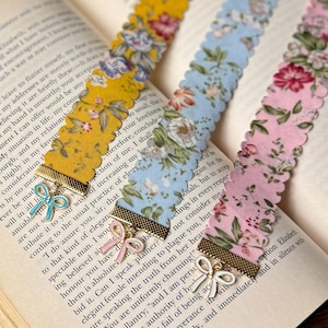 May include: Three floral fabric bookmarks with scalloped edges lie on an open book. The bookmarks are yellow, light blue, and pink, each with a different floral pattern. Each bookmark has a gold-coloured metal accent with a bow charm.
