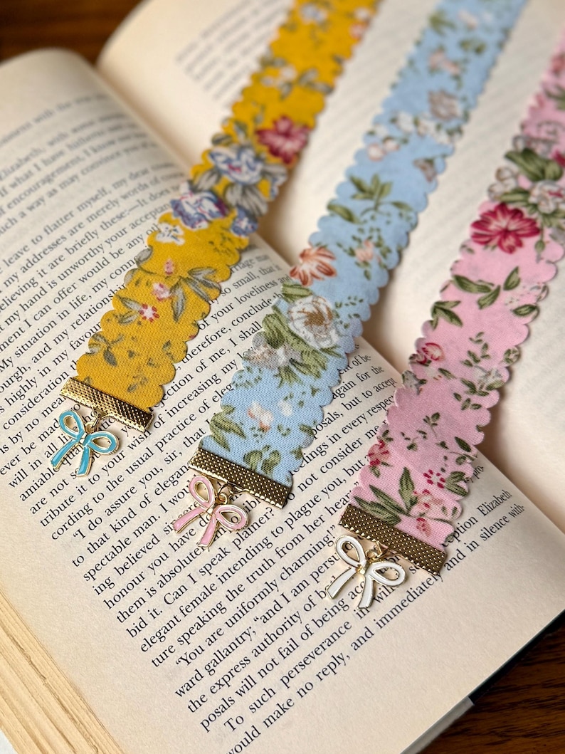 May include: Three floral fabric bookmarks with scalloped edges lie on an open book. The bookmarks are yellow, light blue, and pink, each with a different floral pattern. Each bookmark has a gold-coloured metal accent with a bow charm.