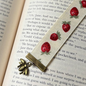 May include: A cream fabric bookmark featuring a strawberry pattern and a bronze bee charm. The bookmark is inserted into a book, with the text of the book visible. The bookmark is approximately 20 cm long.